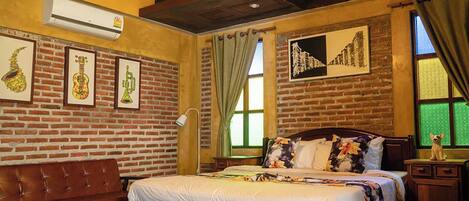 A hotel room with a brick wall, a wooden bed, a desk, and a chair. There is a window with curtains and a picture on the wall.