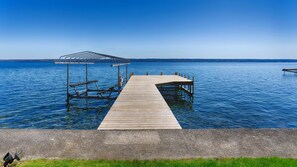 A wooden dock extending into a calm lake, with a covered seating area and a concrete walkway leading to the water.