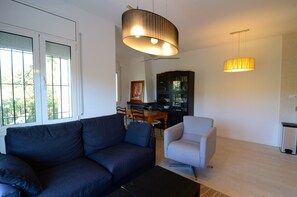 Un salon avec un canapé foncé, un fauteuil clair, une table à manger avec des chaises, une bibliothèque et un grand luminaire circulaire.