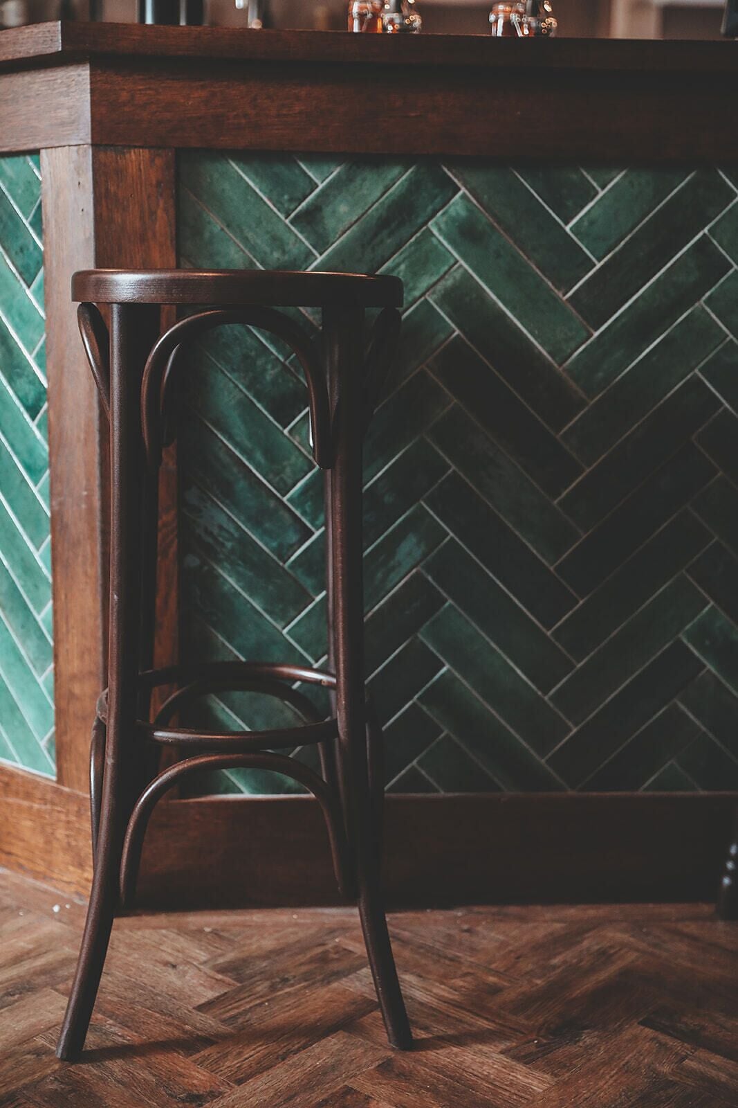 A bar stool in a room with a herringbone-patterned green tile backsplash and wooden flooring.