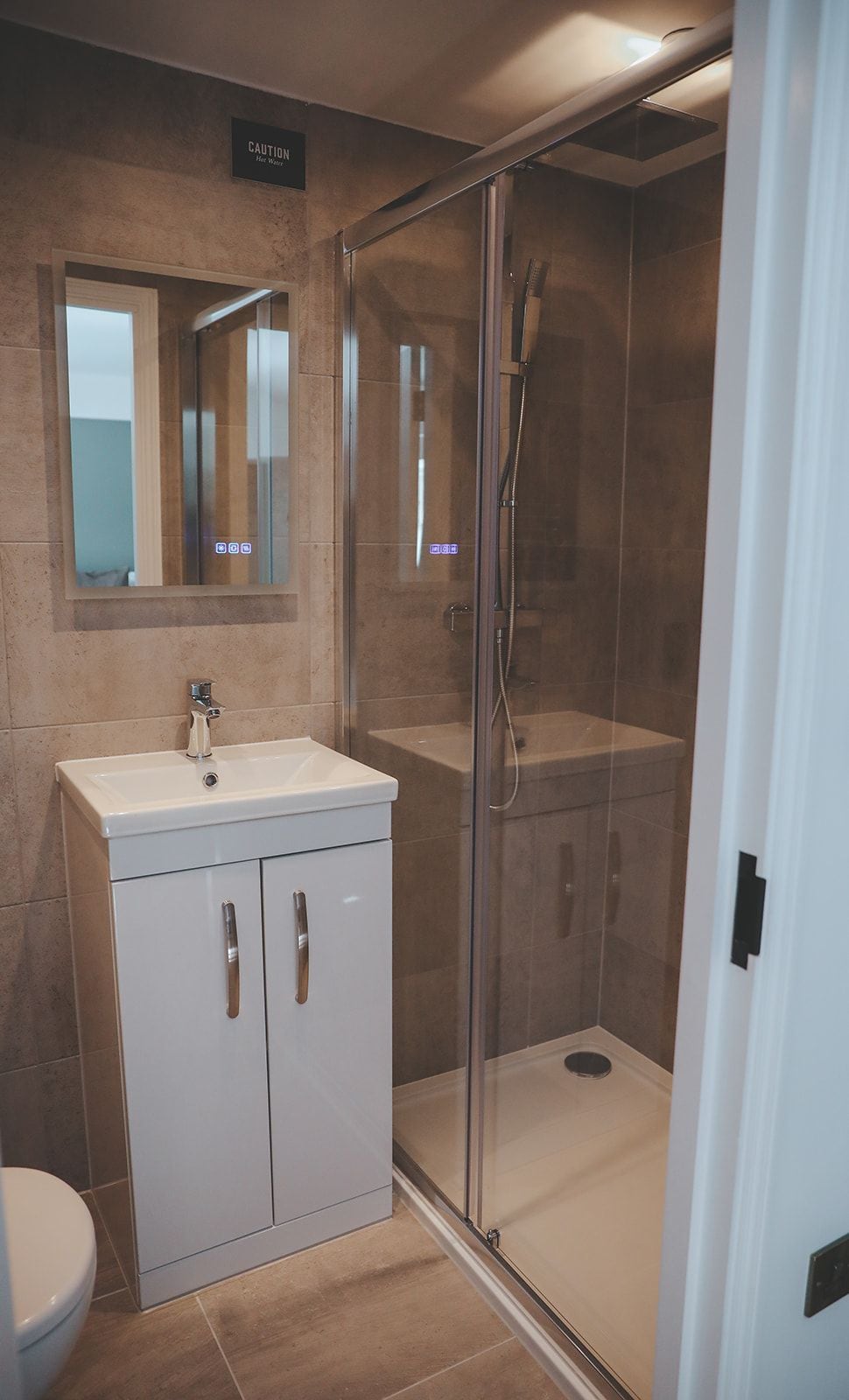 A bathroom with a glass-enclosed shower, a sink with a mirror above it, and a caution sign on the wall.