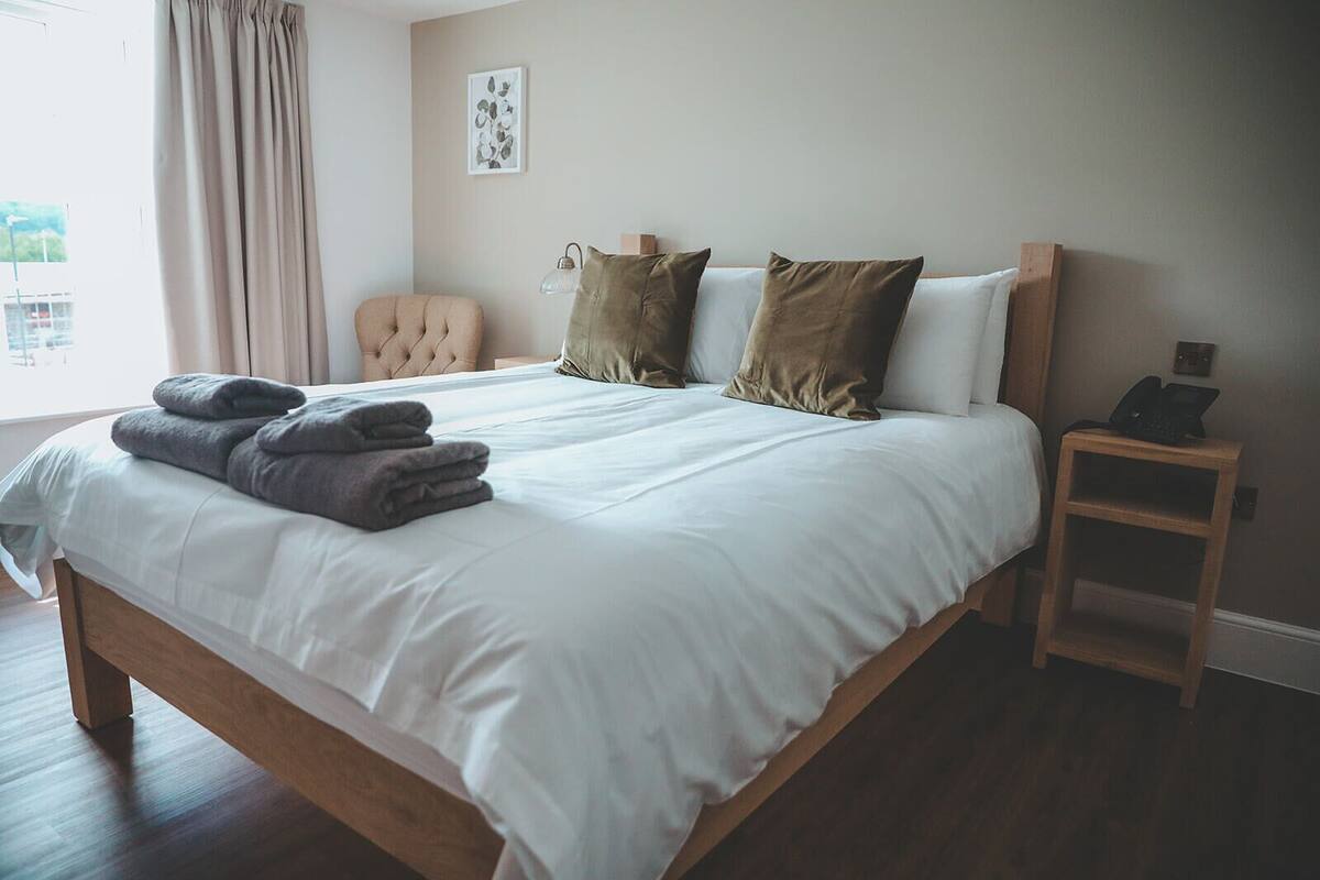 A neatly made bed with white linens and two pillows, a bedside table with a lamp, and a chair in a room with a window and a wall-mounted picture.