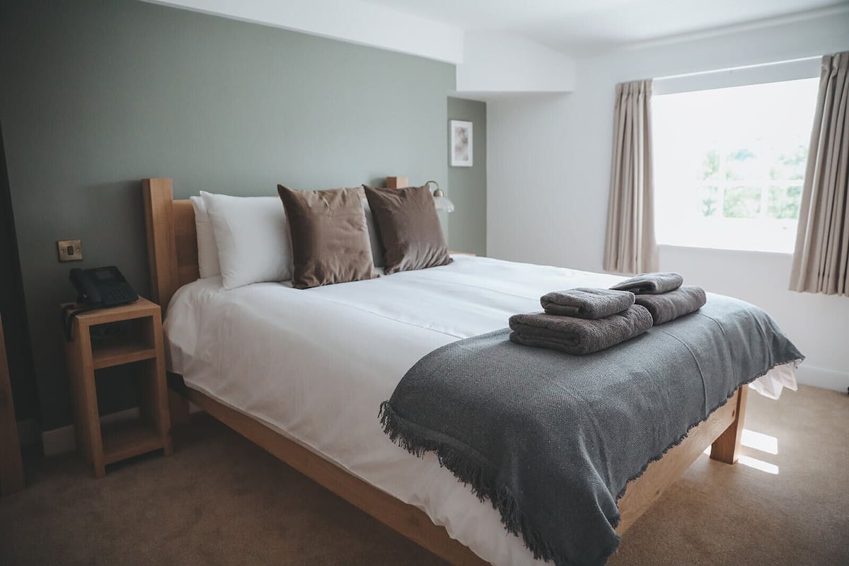 A neatly made bed with a grey blanket and pillows, a bedside table with a phone, and a window with curtains.