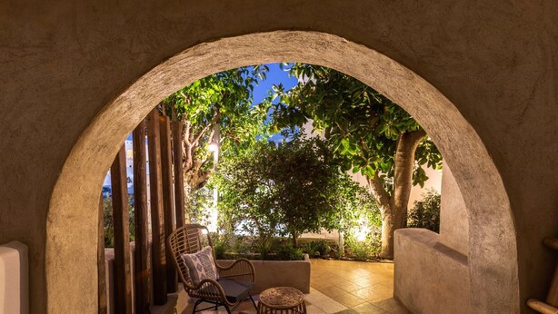 Un área de estar al aire libre con un arco de piedra, una silla de mimbre, un banco acolchado y un taburete tejido.