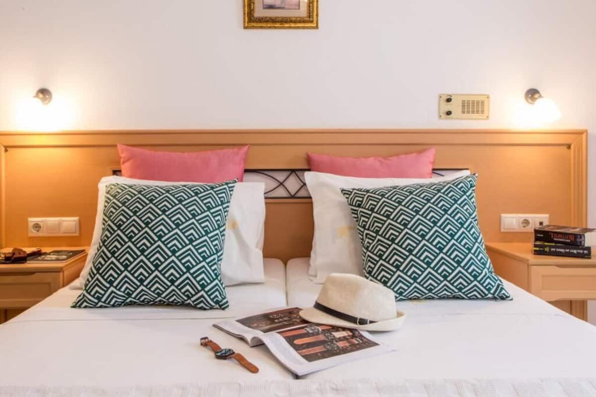 Una cama bien hecha con almohadones estampados, un sombrero y un libro sobre una colcha blanca.