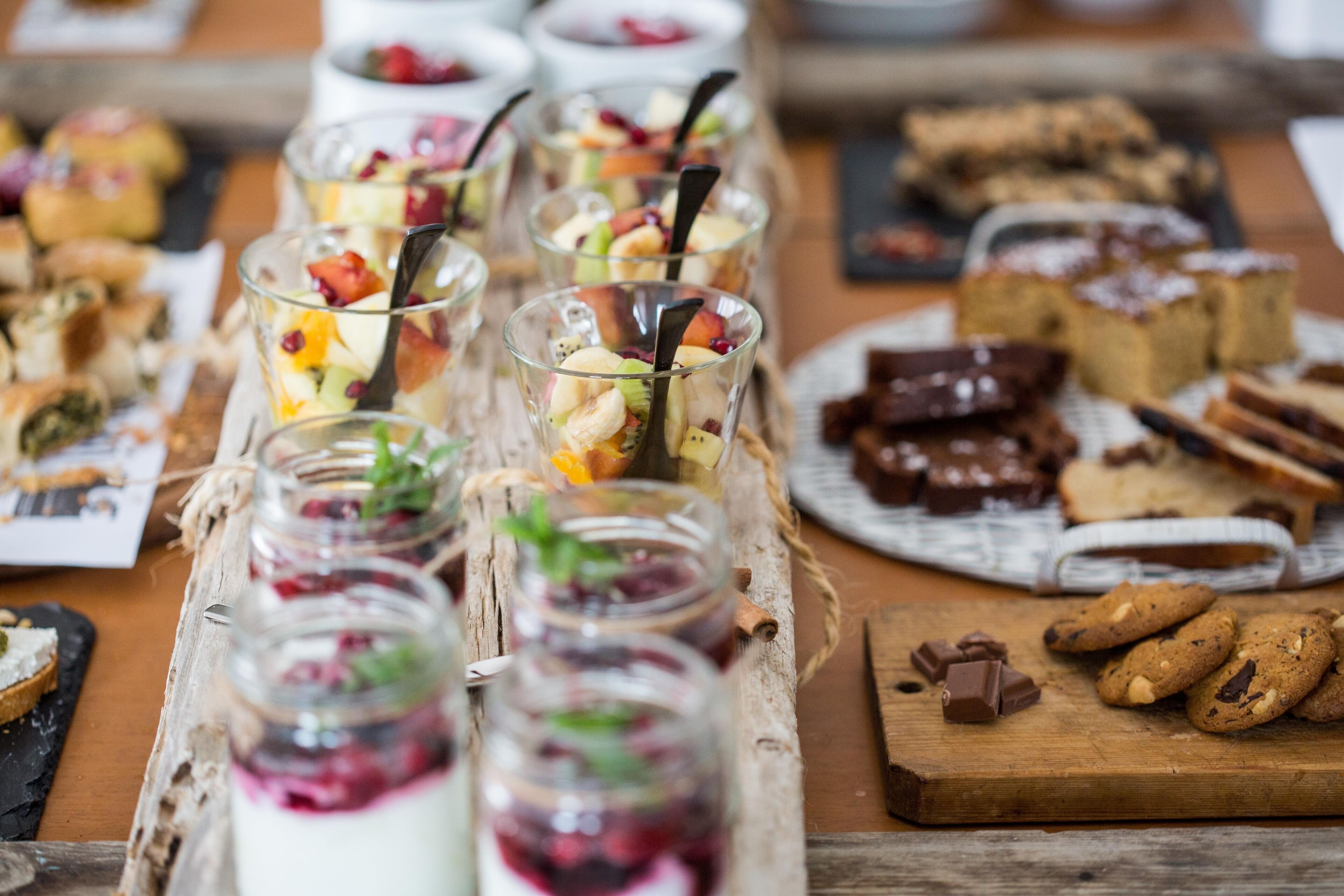 Diversos postres y bocadillos dispuestos sobre una mesa de madera, entre ellos, frascos con fruta y crema, galletas y otras preparaciones horneadas.