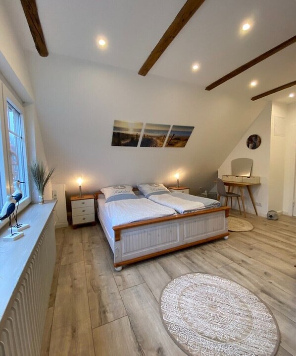 A bedroom with a sloped ceiling, wooden beams, a bed with white bedding, a desk with a chair, and a small round rug.