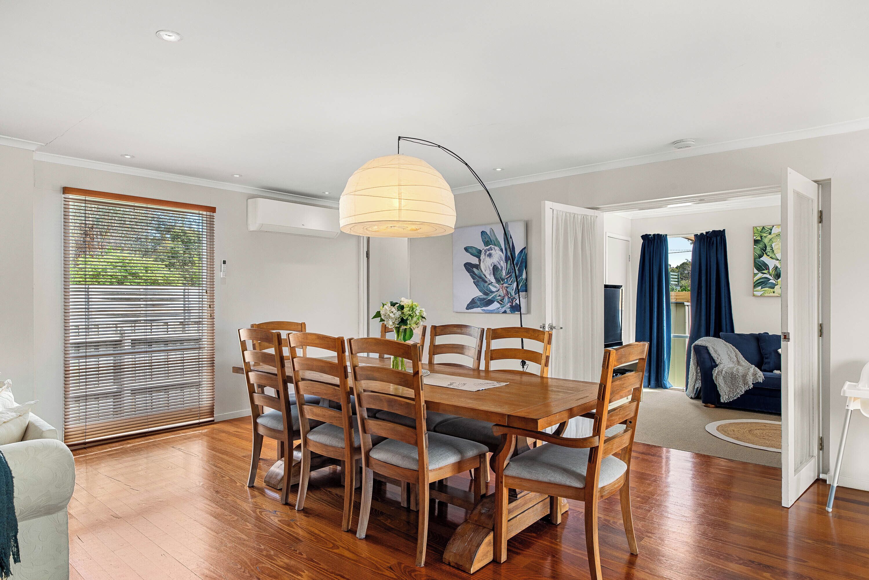 A dining area with a wooden table and chairs, a hanging light fixture, and a view of the outdoors through a window with blinds.