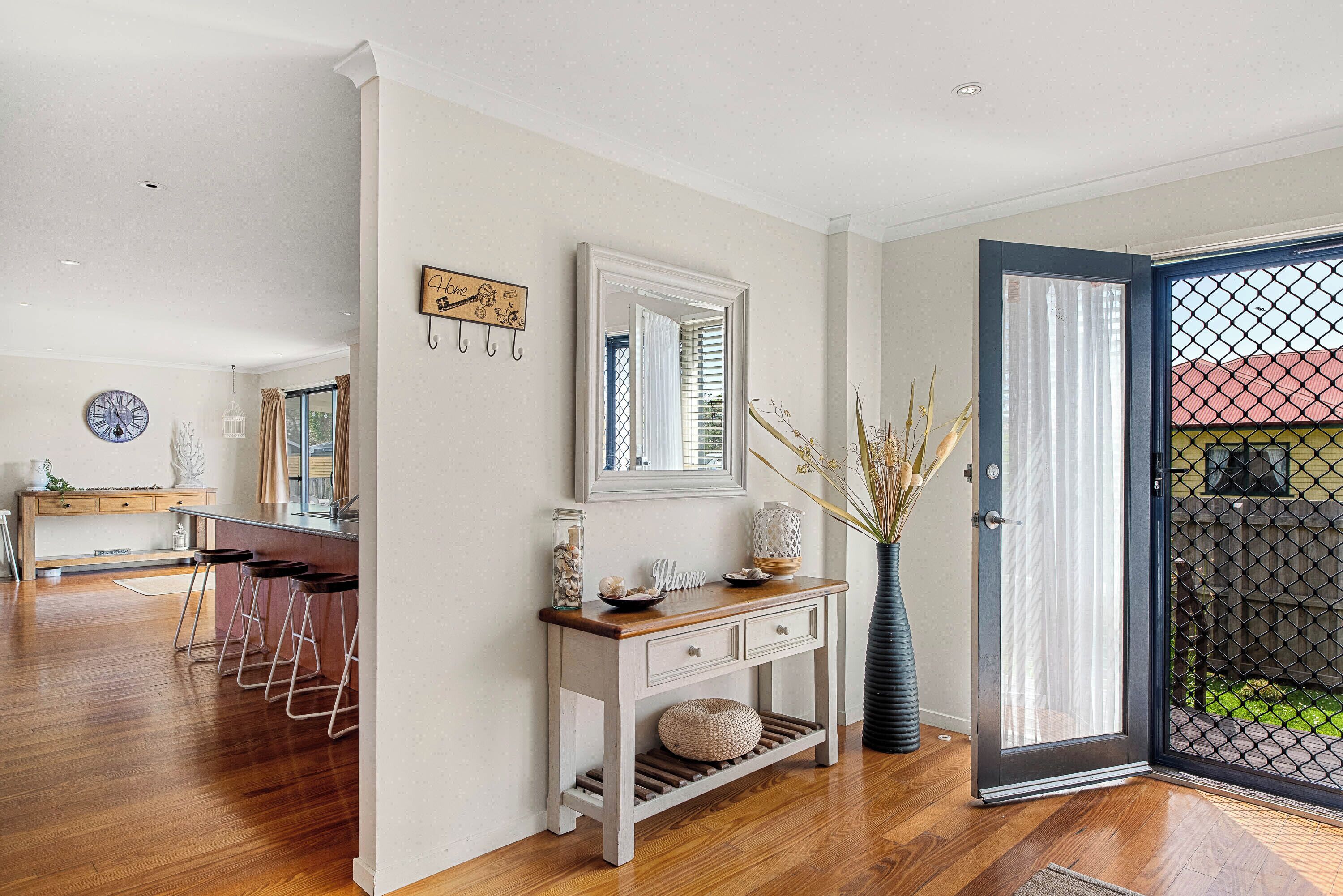 Une cuisine moderne avec un plancher en bois, un coin repas avec des tabourets de bar, une console blanche agrémentée d’objets décoratifs et un grand miroir au-dessus.