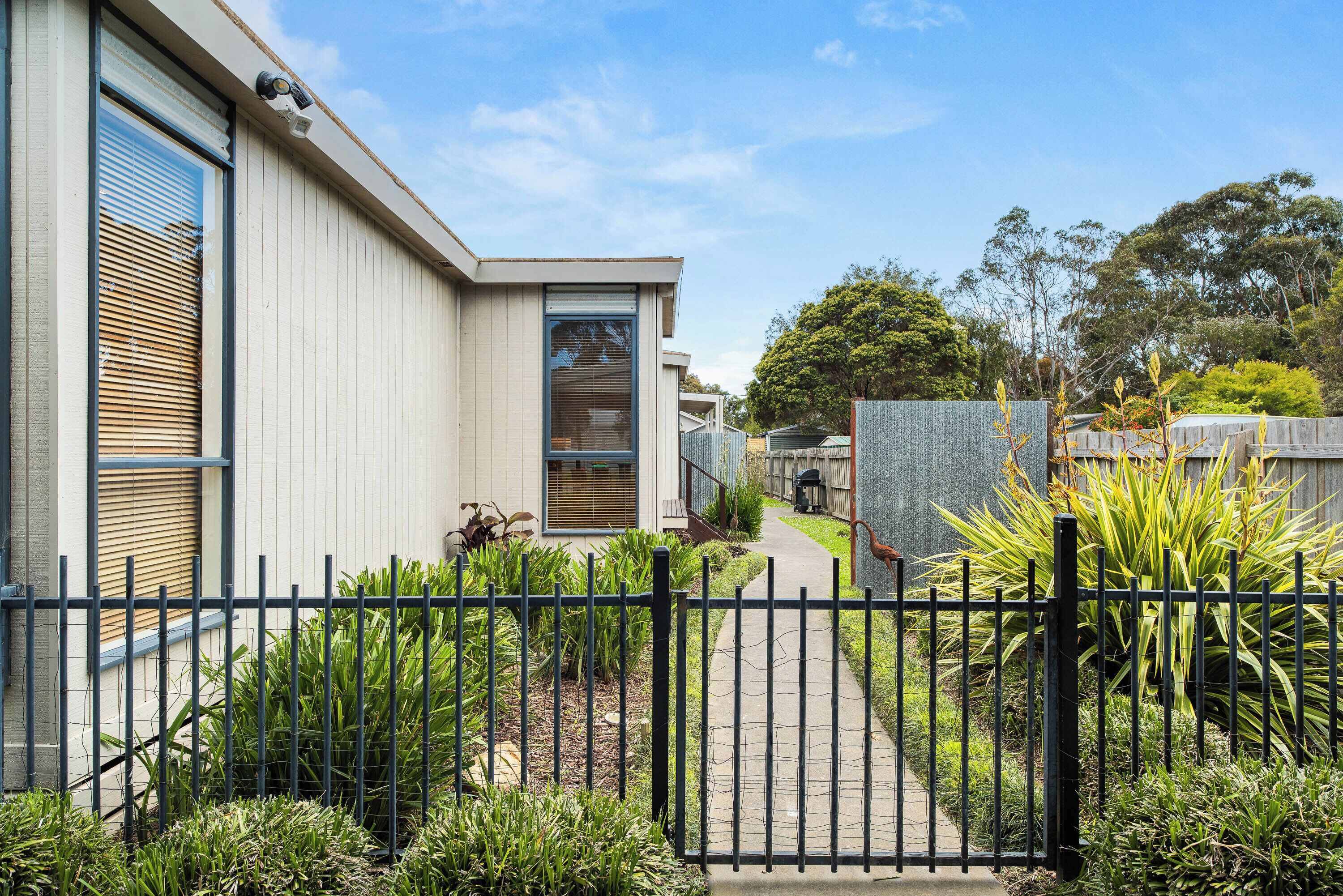 A modern building with a gated entrance, a pathway leading to the main door, and a well-maintained garden with various plants.