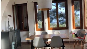 A dining area with a wooden table, four chairs, a dark-colored cabinet, and large windows with a view of trees and hills. - Maison en Pierres Avec vue sur mer et les Îles (Bormes-les-Mimosas)