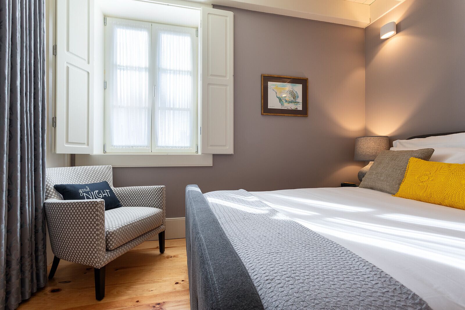 A bedroom with a bed, a chair, a window with curtains, a wall-mounted lamp, and a framed picture.
