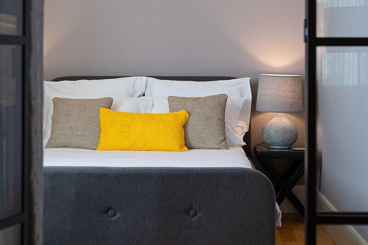 A bedroom with a bed, pillows, a lamp, and a window.