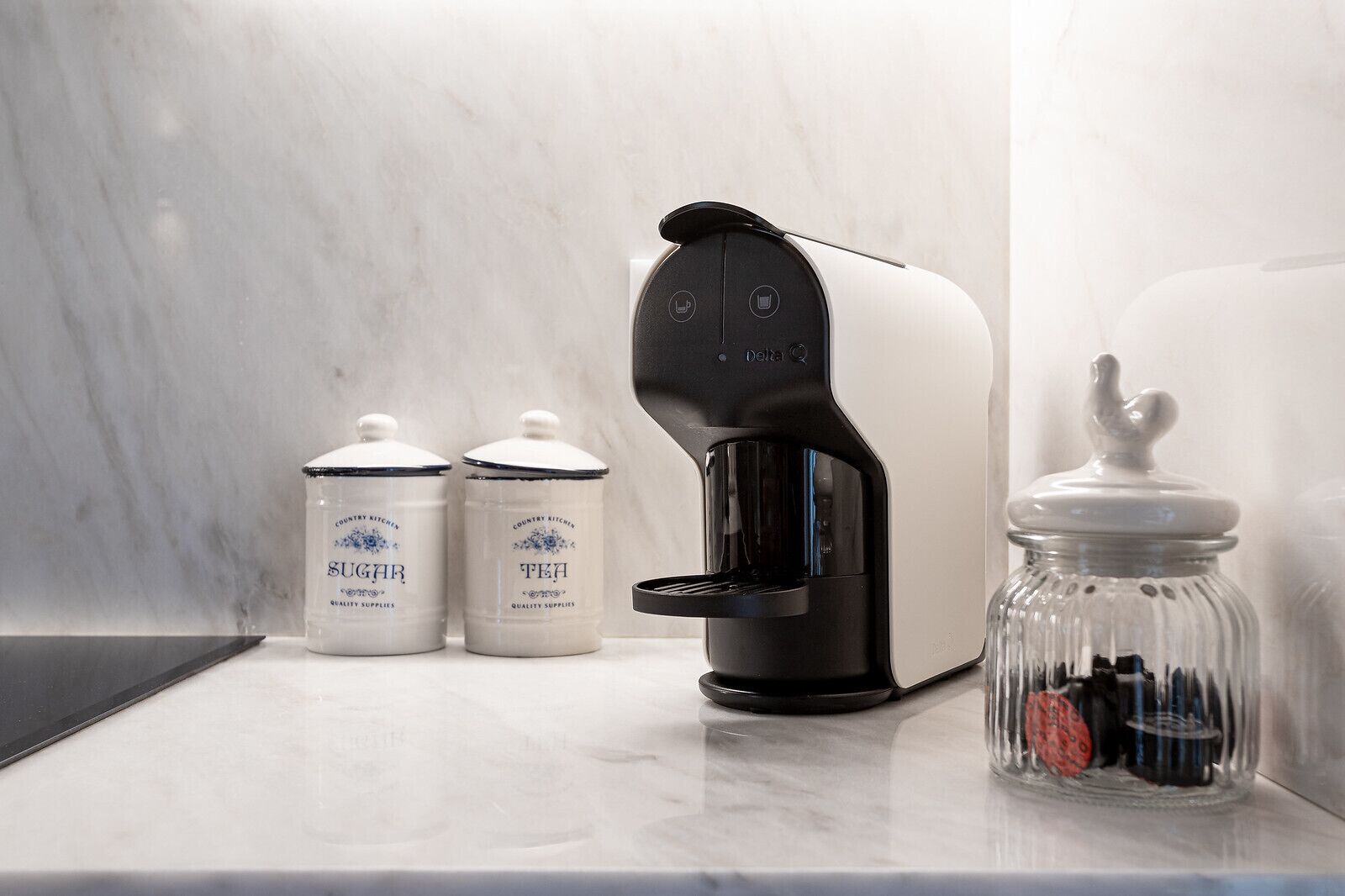 A coffee maker on a marble countertop with two jars labeled "SUGAR" and "TEA" and a glass jar with a lid.