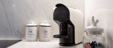 A coffee maker on a marble countertop with two jars labeled "SUGAR" and "TEA" and a glass jar with a lid.