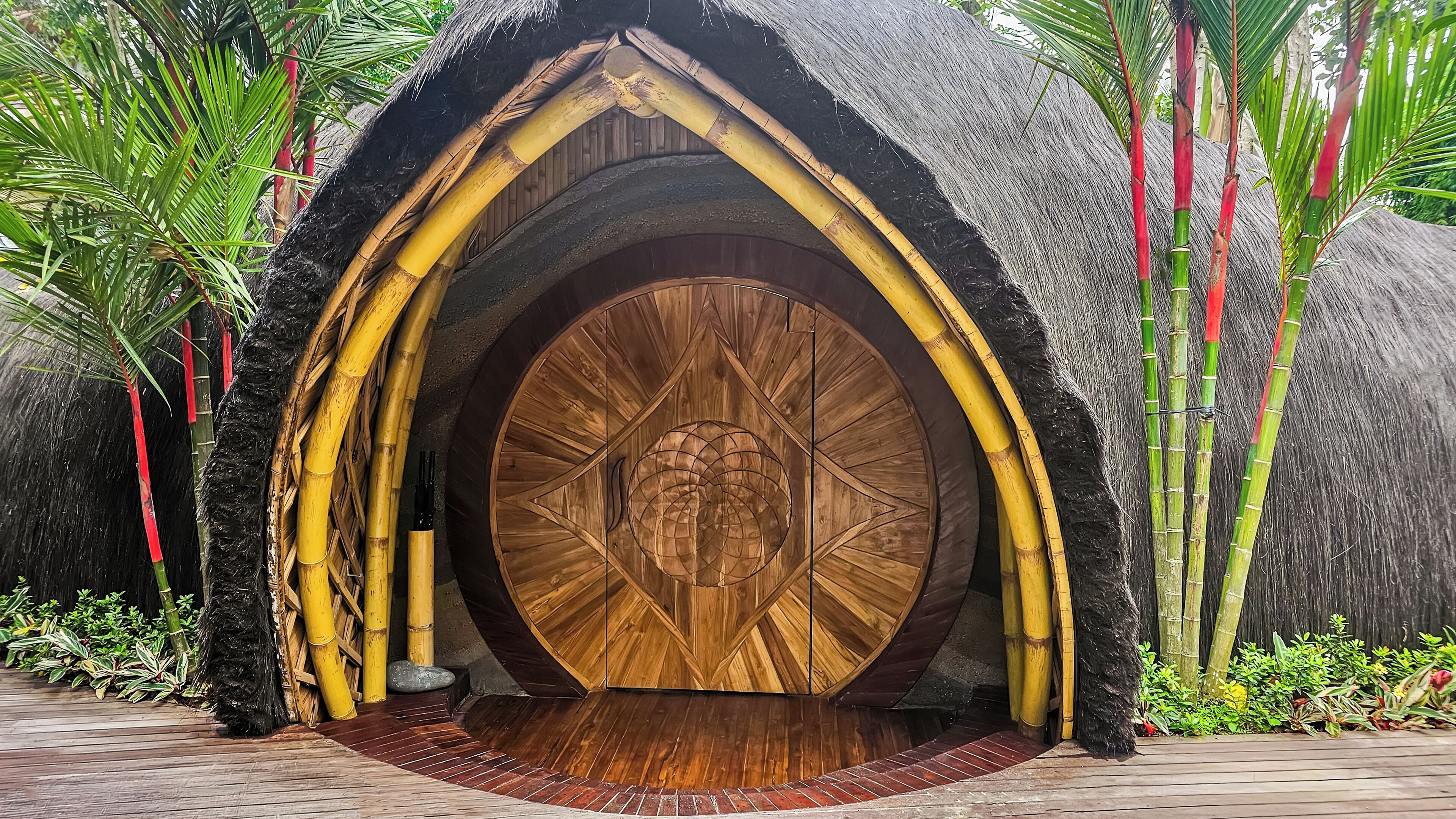 Una puerta de madera grande con un diseño circular, rodeada de bambú y palmeras.