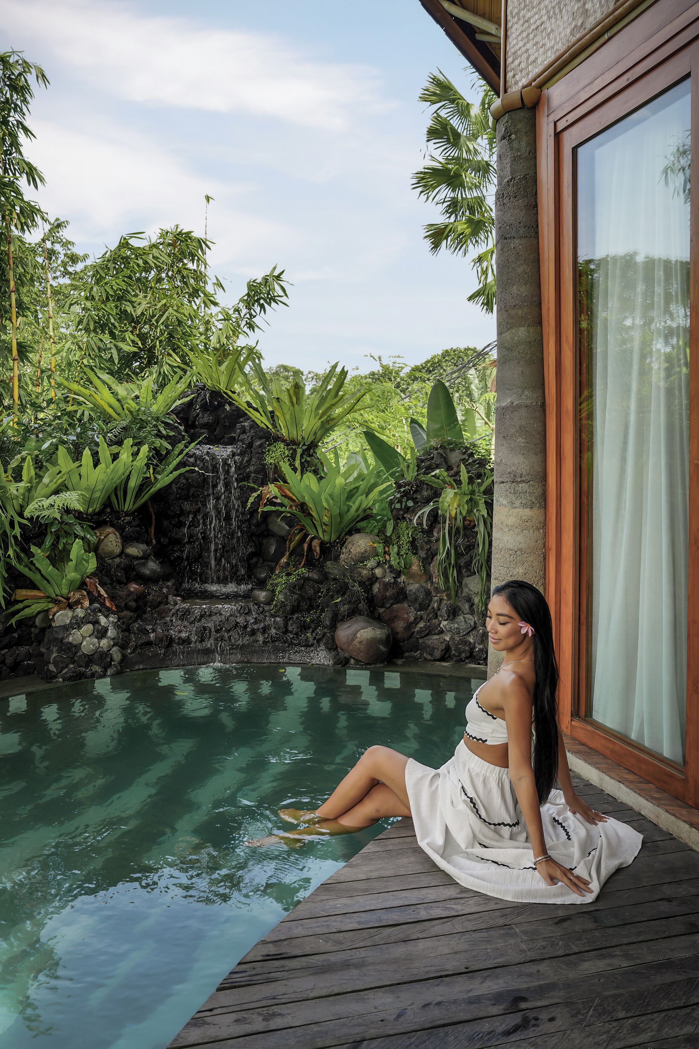 A person sitting by a pool with a waterfall and tropical plants in the background.