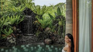 A person sitting by a pool with a waterfall and tropical plants in the background.