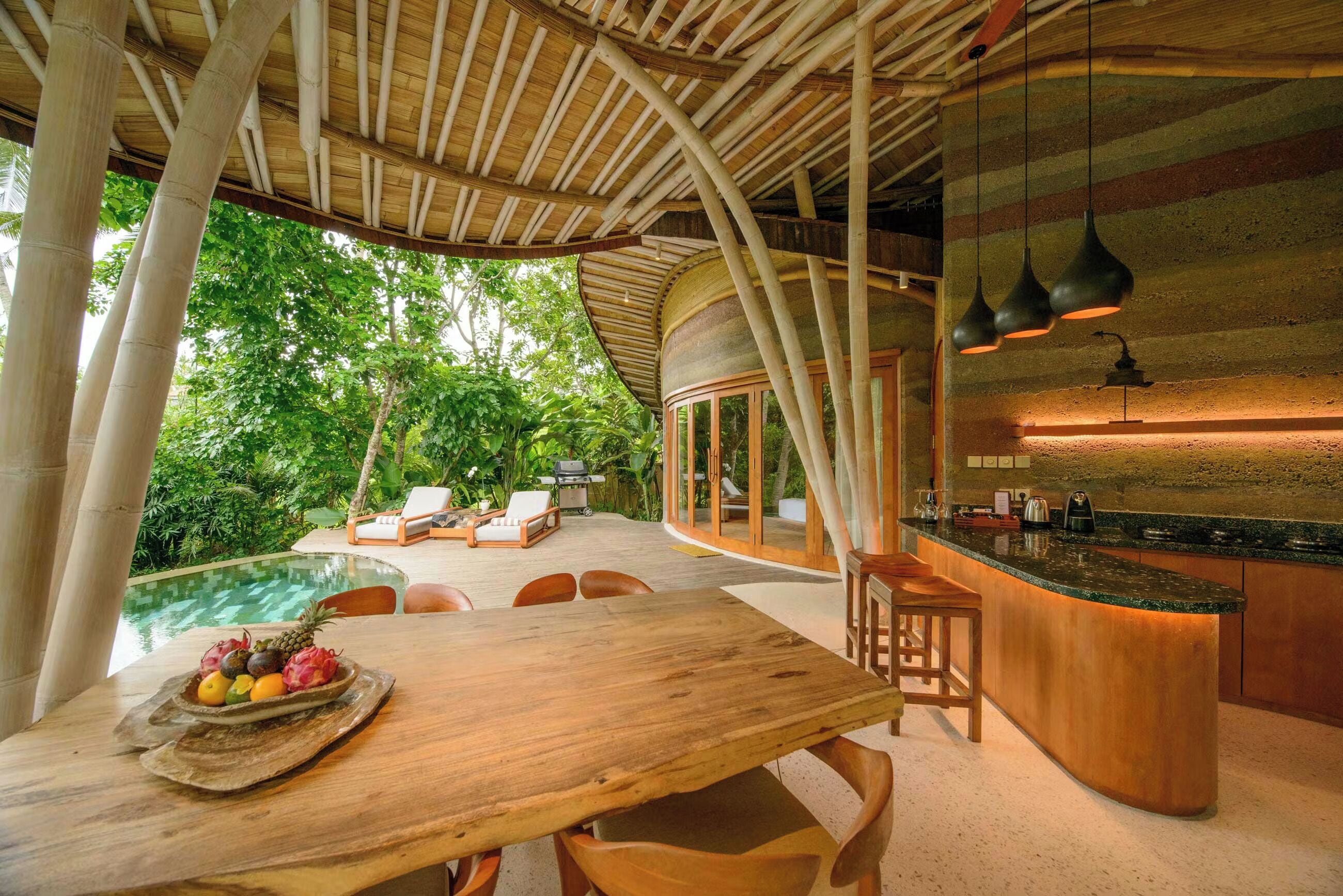 A wooden dining table with fruit, a pool, and a kitchen area with a curved island.