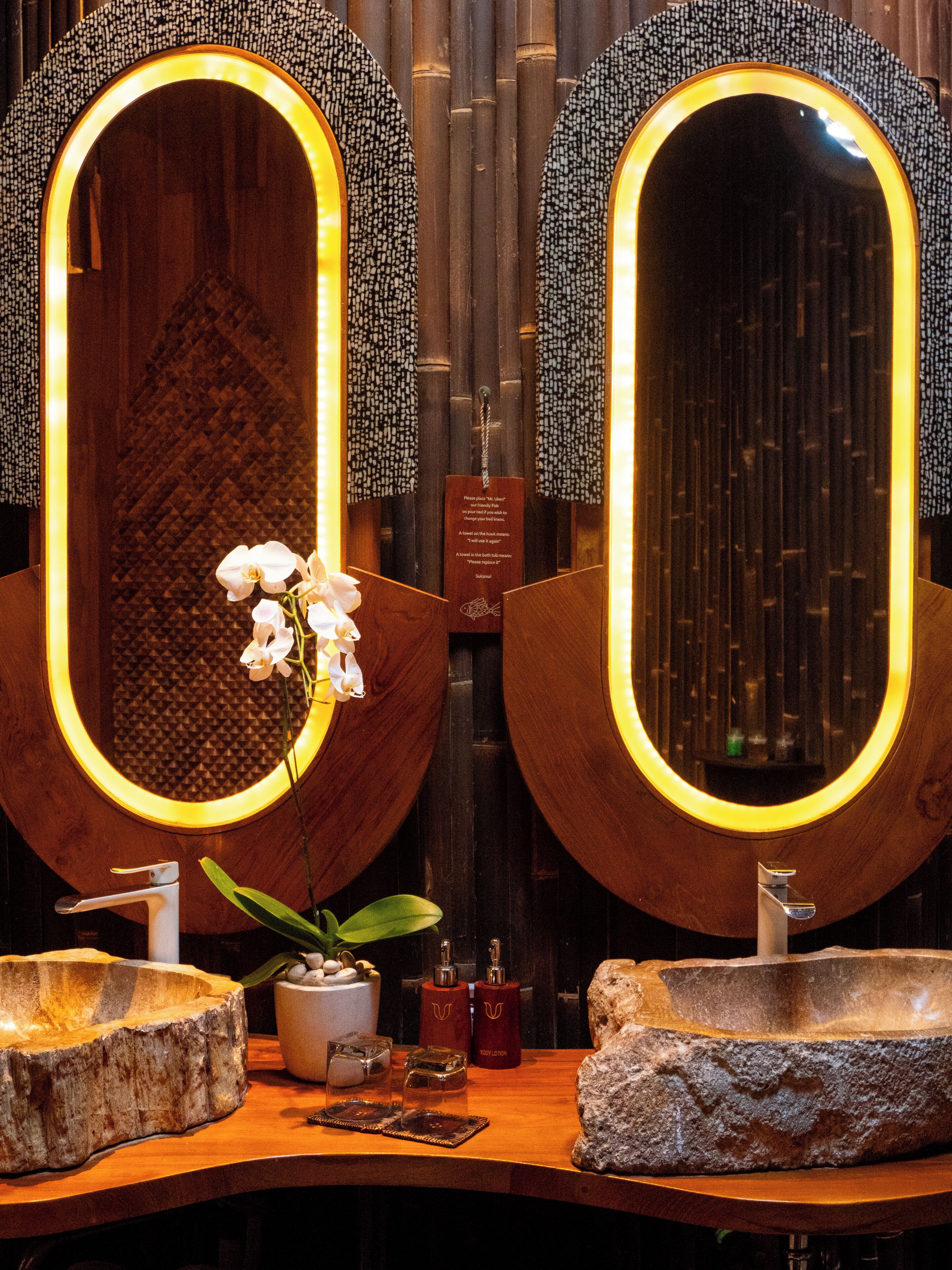 Two oval mirrors with illuminated edges, a wooden vanity with a sink, a small plant, and a plaque on the wall.