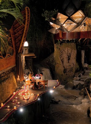 A dining area with a waterfall backdrop, wooden structures, and a carved stone wall. -  Ulaman Eco Luxury Resort (Tabanan)