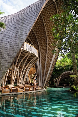 A poolside area with a unique architectural design, including a curved roof and a wooden deck with lounge chairs. -  Ulaman Eco Luxury Resort (Tabanan)