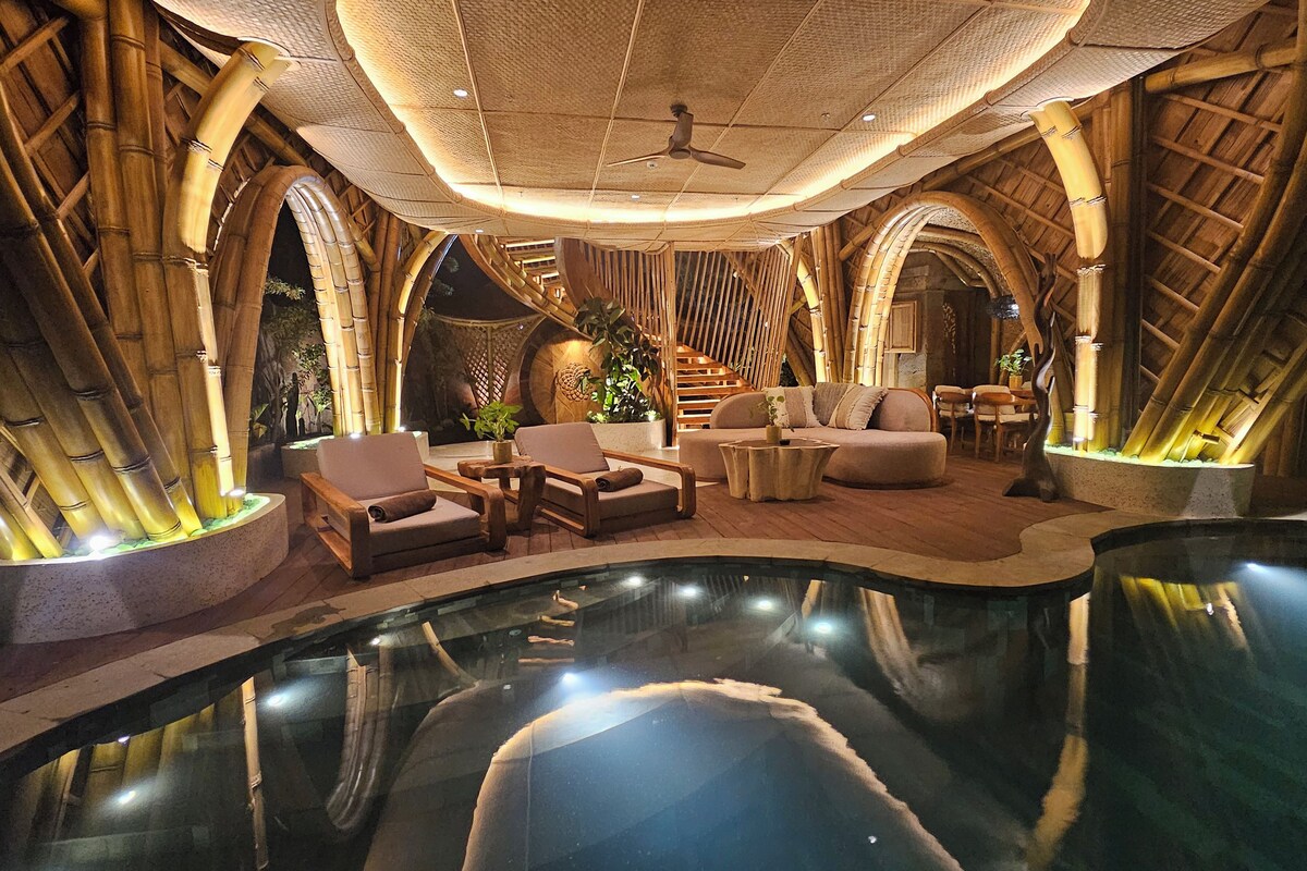 A luxurious indoor pool area with bamboo architecture, seating arrangements, and a ceiling fan.