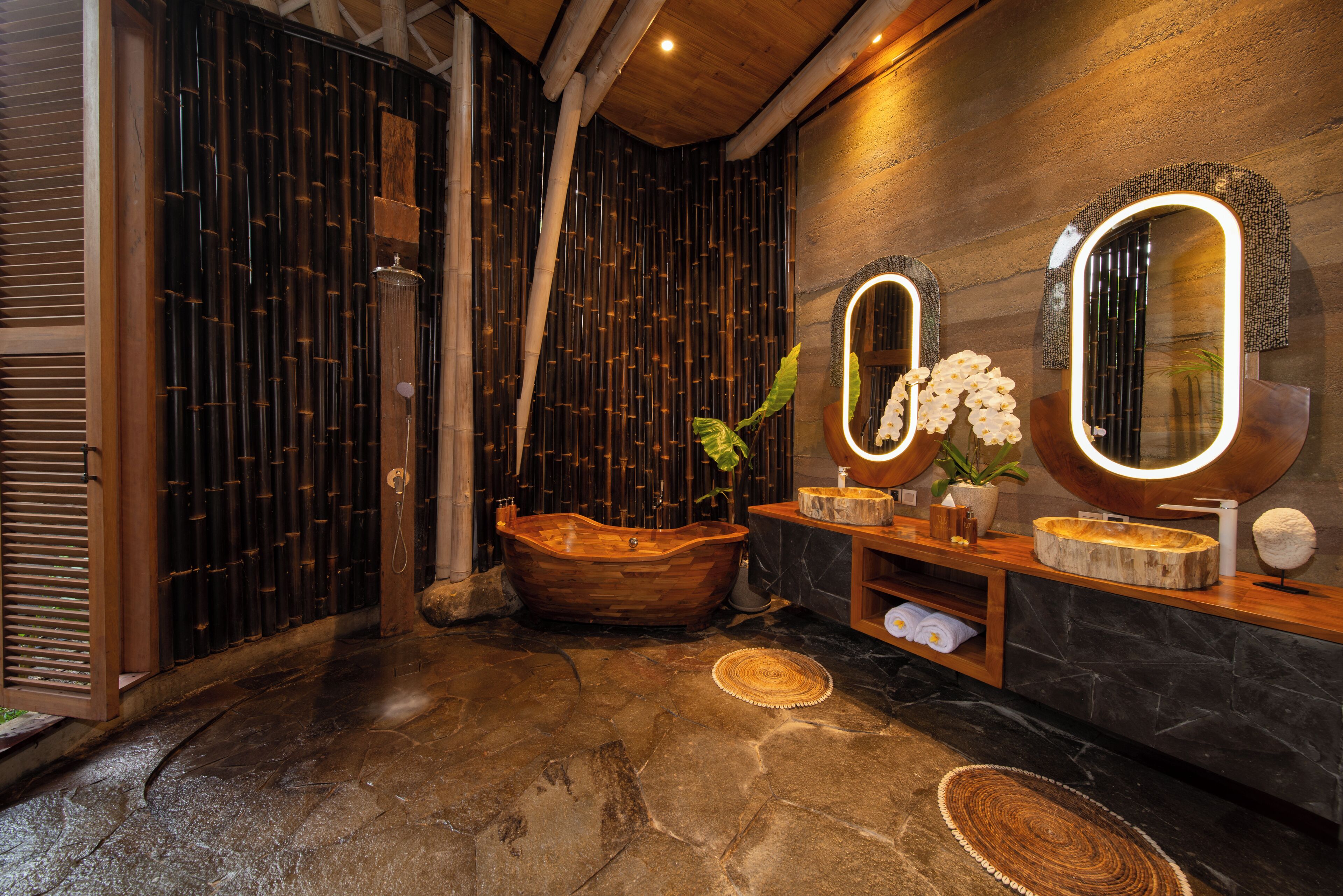 Une salle de bain avec une baignoire en bois, deux miroirs éclairés, un sol en pierre et un mur en bambou.