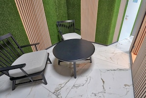 Pequeño espacio al aire libre amurallado con una mesa redonda y dos sillas, rodeado de listones de madera verticales y césped artificial.