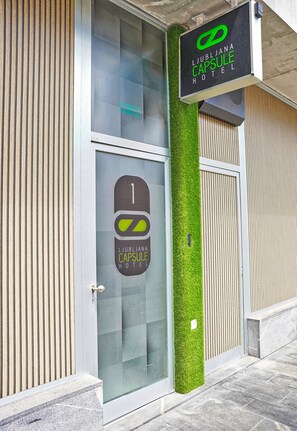 Una entrada de hotel moderna con puerta de vidrio, un jardín vertical verde y un letrero que dice Lujbjana Capsule Hotel.