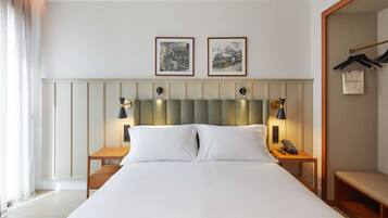 A neatly made bed with white linens, two bedside tables with lamps, a wooden bench, and a coat rack with a hanging bag.