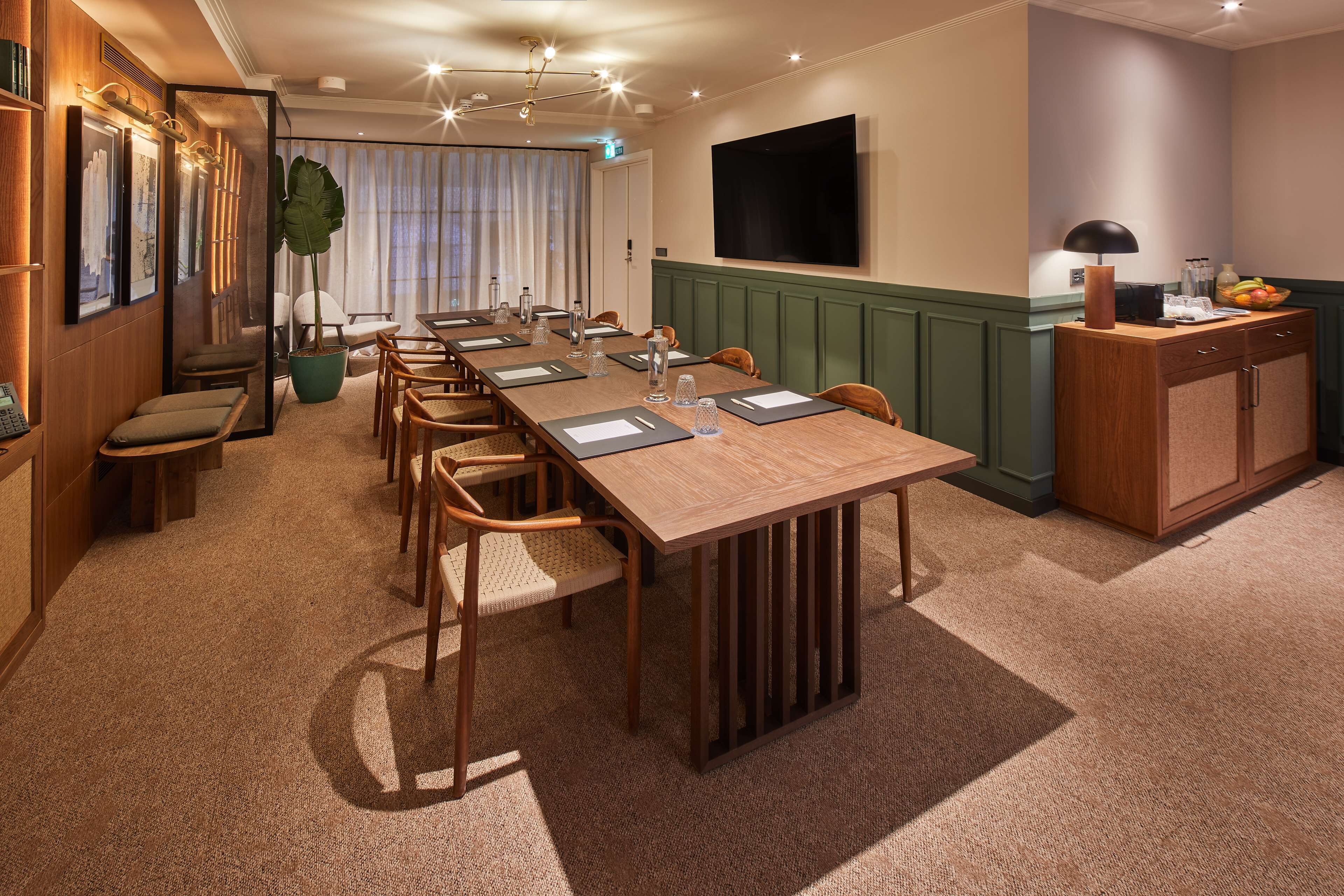 Un salón de conferencias con una larga mesa, sillas, una planta grande, un televisor y un mueble con fruta.