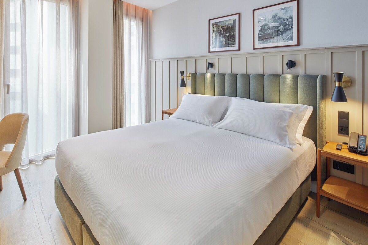 Ein Hotelzimmer mit einem großen Bett, einem Nachttisch mit Lampe, einem Sessel und zwei gerahmten Bildern an der Wand.