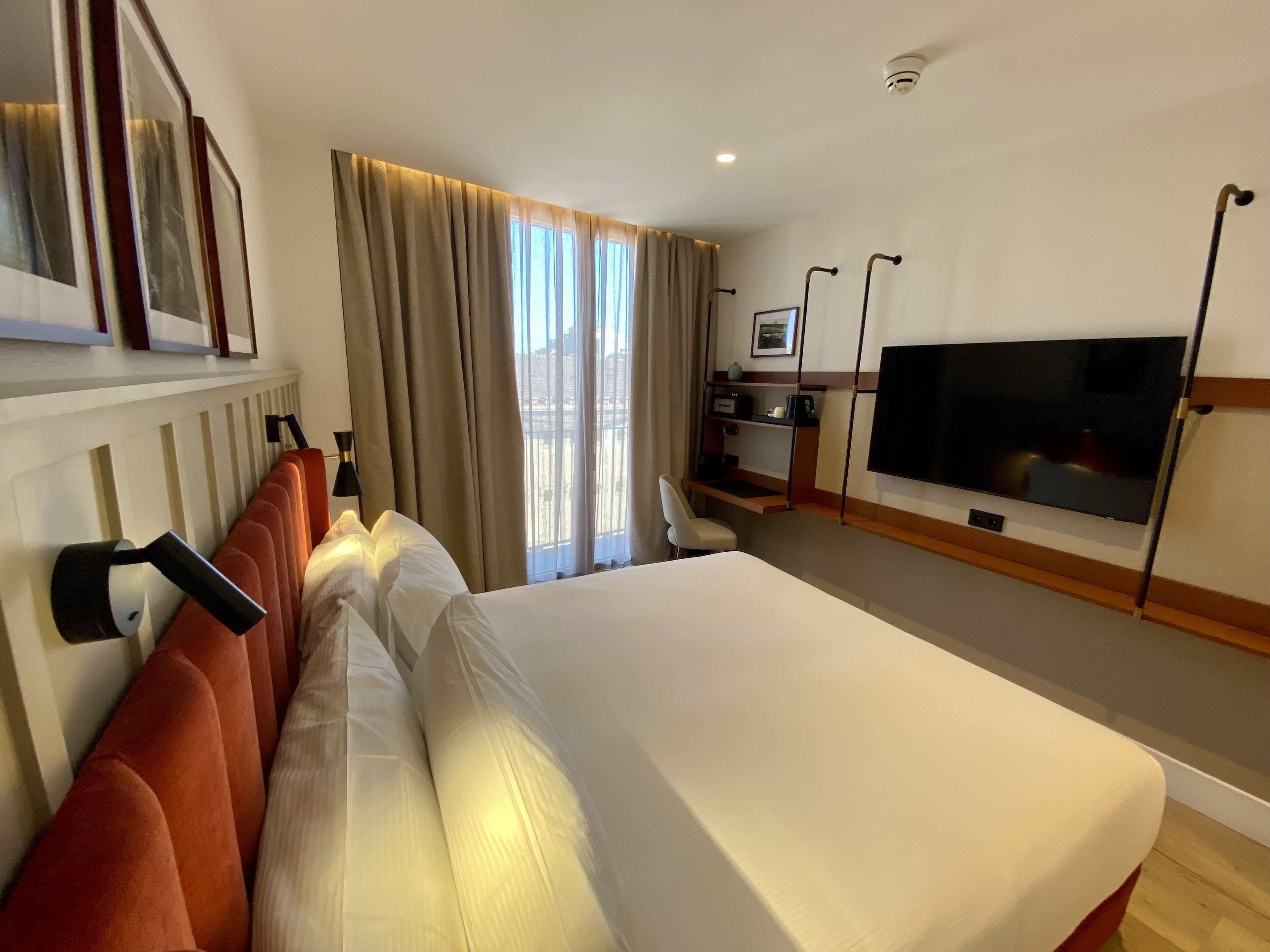Una habitación de hotel con una cama grande, televisión, escritorio con computadora y ventana con cortinas.