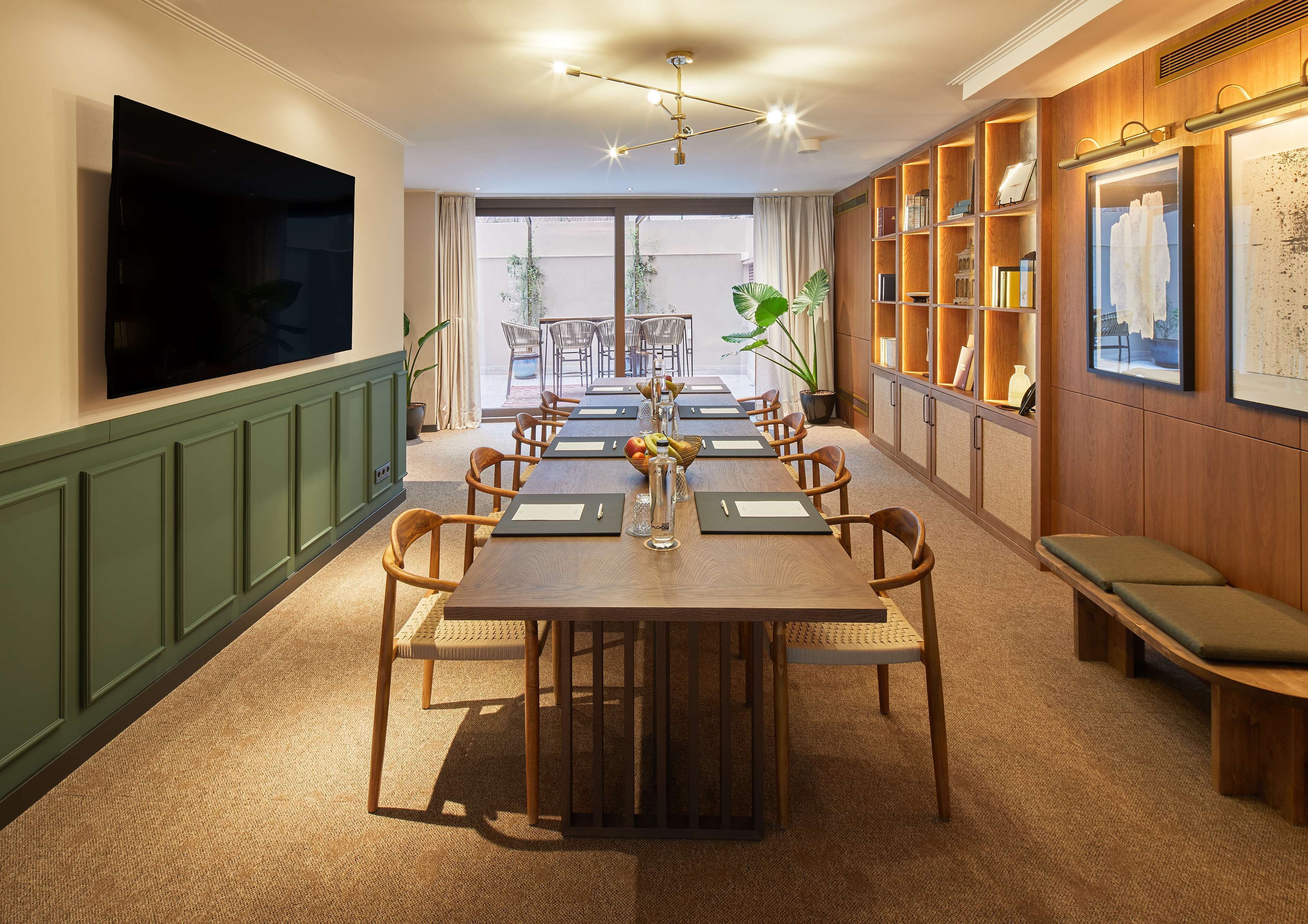 A dining area with a long table, chairs, a TV mounted on the wall, and a bookshelf with framed pictures.