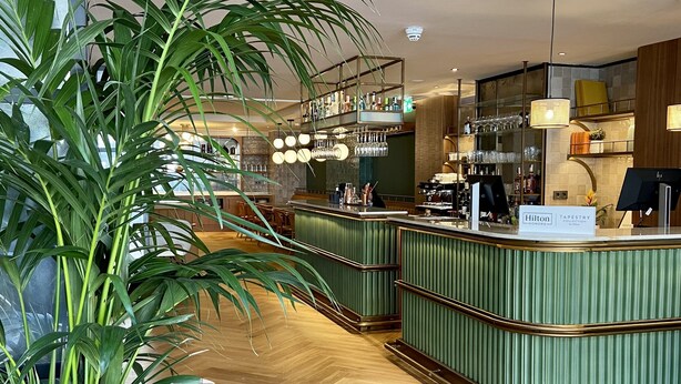 A modern hotel lobby with a curved reception desk, a large potted plant, and a bar area with hanging lights. - Via Sants Hotel Barcelona, Tapestry Collection by Hilton (Barcelona)