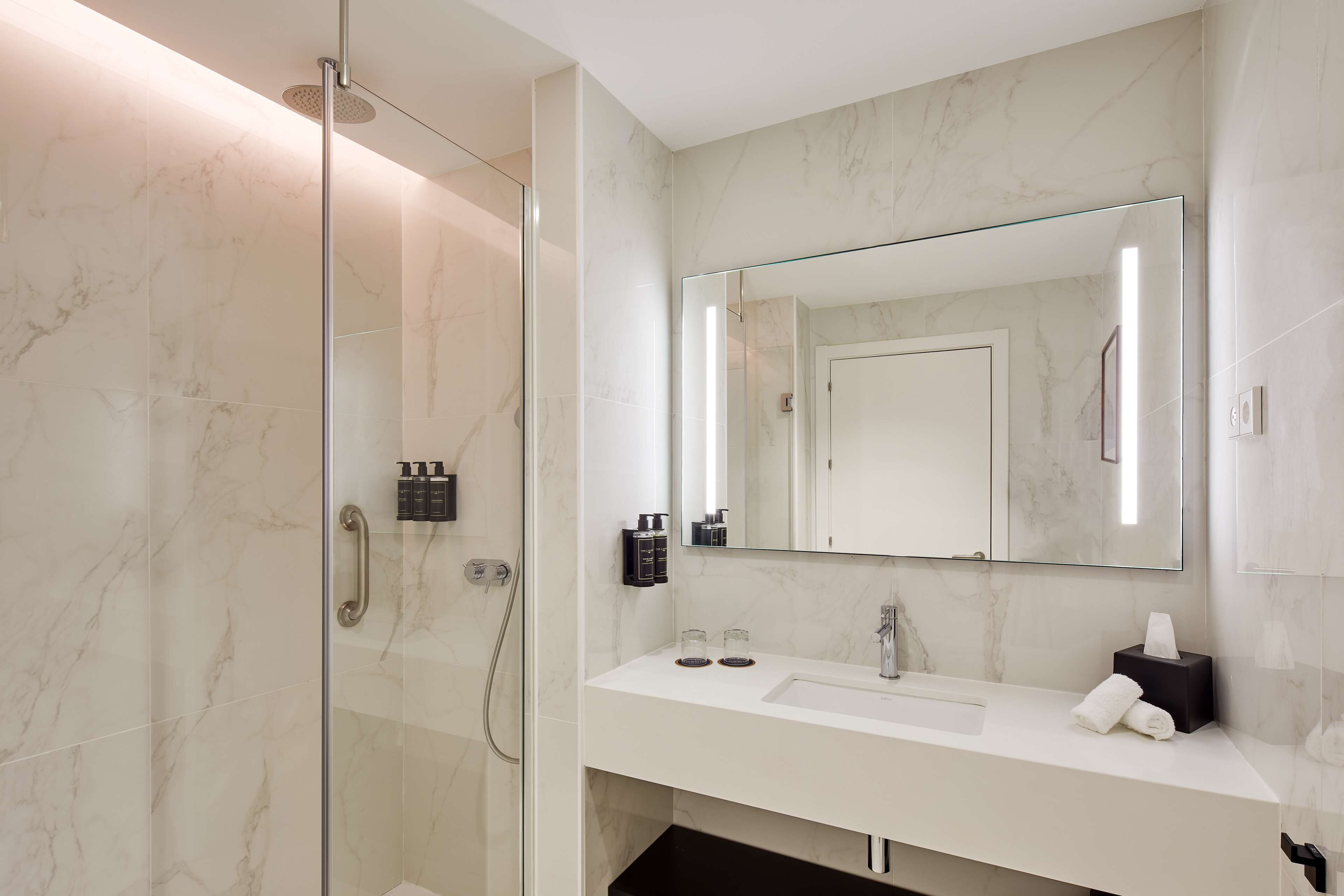 A modern bathroom with a glass-enclosed shower, a large mirror, and a white vanity with a sink and towel dispenser.
