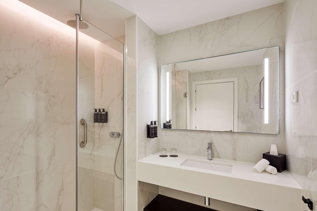A modern bathroom with a glass shower enclosure, a white sink, a large mirror, and marble walls.
