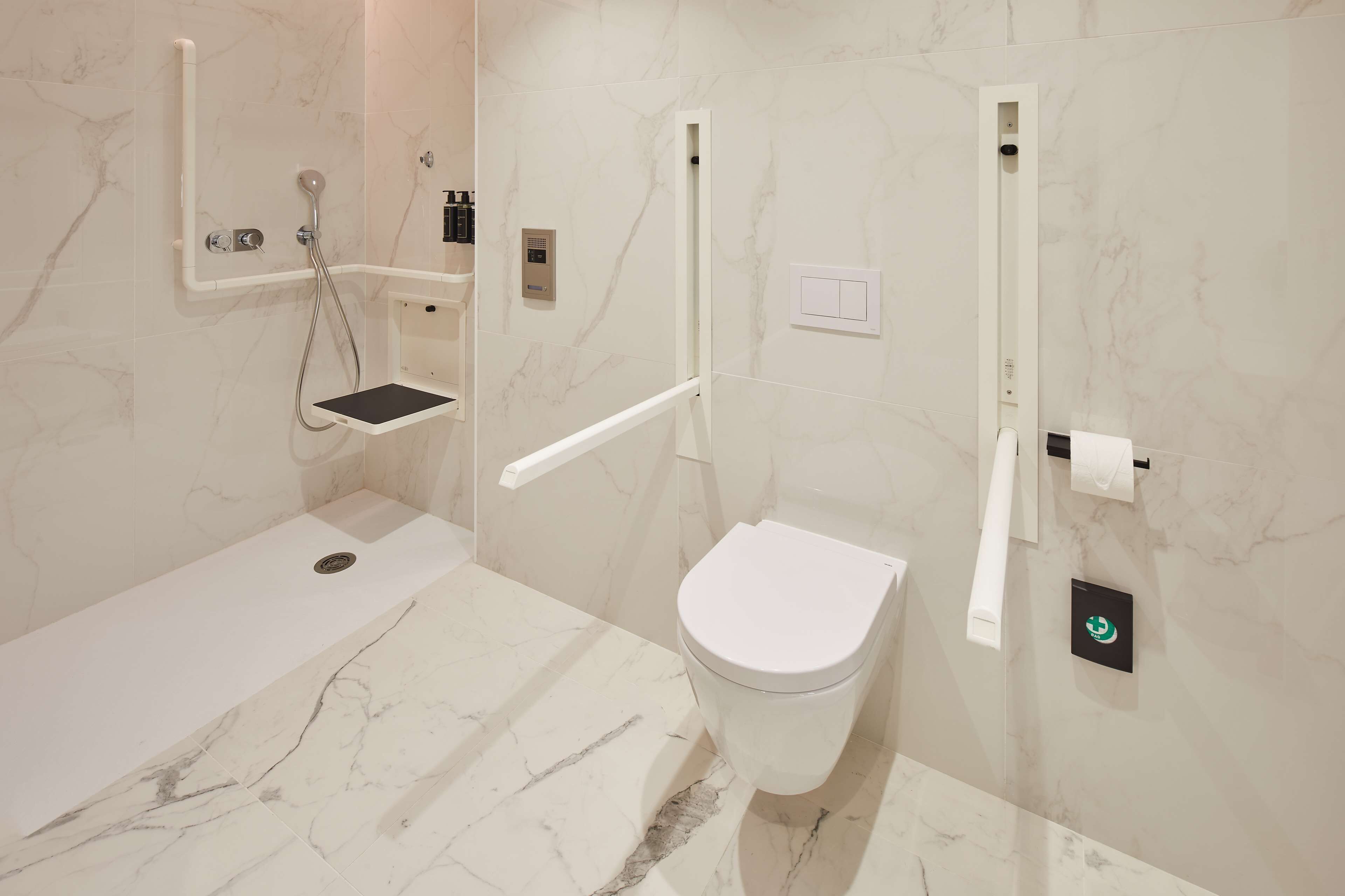 Un baño moderno con un inodoro blanco, una zona de ducha con barra de apoyo y suelo de mármol.