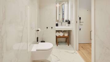 Een moderne badkamer met een wit toilet, een wastafel en een spiegel. Er is een houten plank met handdoeken en een klein tafeltje.