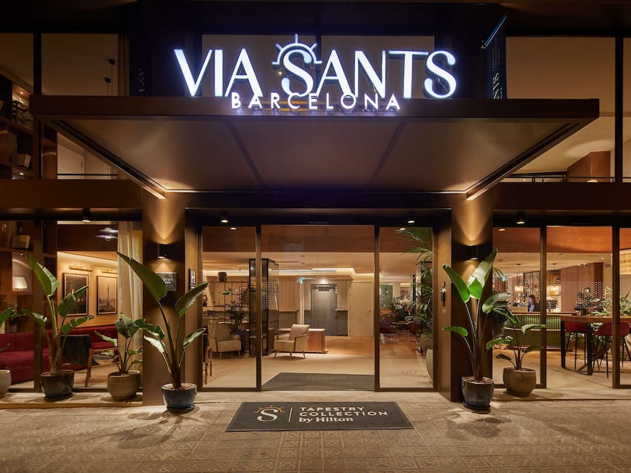 The entrance of Via Sants Barcelona with a modern design, featuring a well-lit facade, potted plants, and a reception area visible through the glass doors.