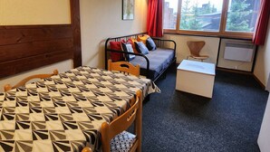 A small, cozy room with a bed, a desk, and a chair. There is a window with curtains, a radiator, and a picture on the wall.