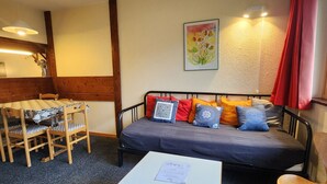 A cozy room with a bed, a small table, and a chair. There's a painting on the wall and a lamp on the side.