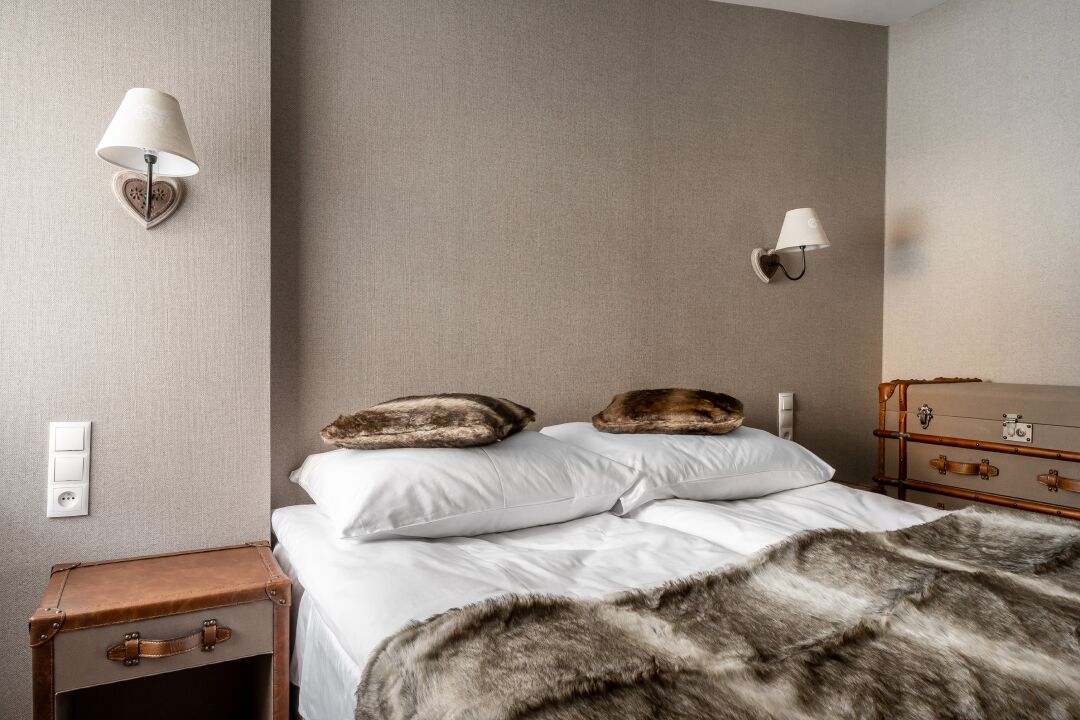 A bedroom with a bed, two wall-mounted lamps, a suitcase, and a fur throw.