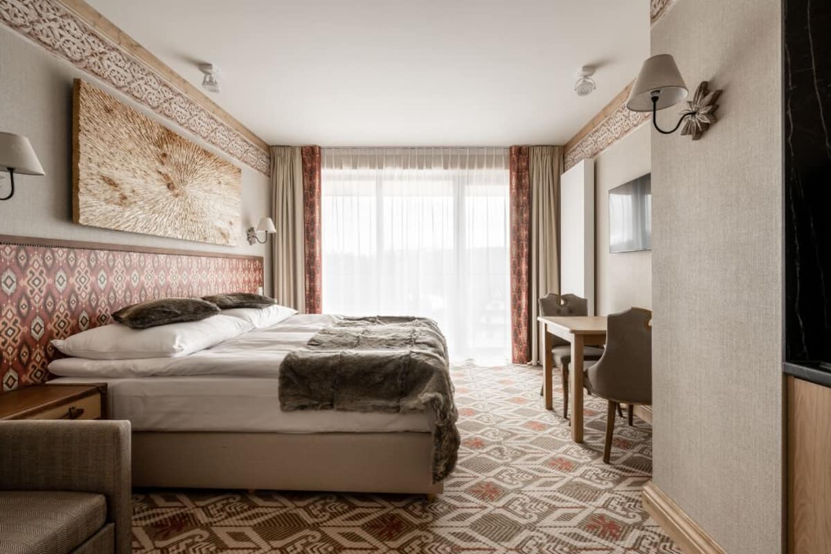 Une chambre d’hôtel avec un grand lit, un bureau et une chaise. On y trouve une fenêtre avec des rideaux, une lampe fixée au mur et un élément décoratif mural.