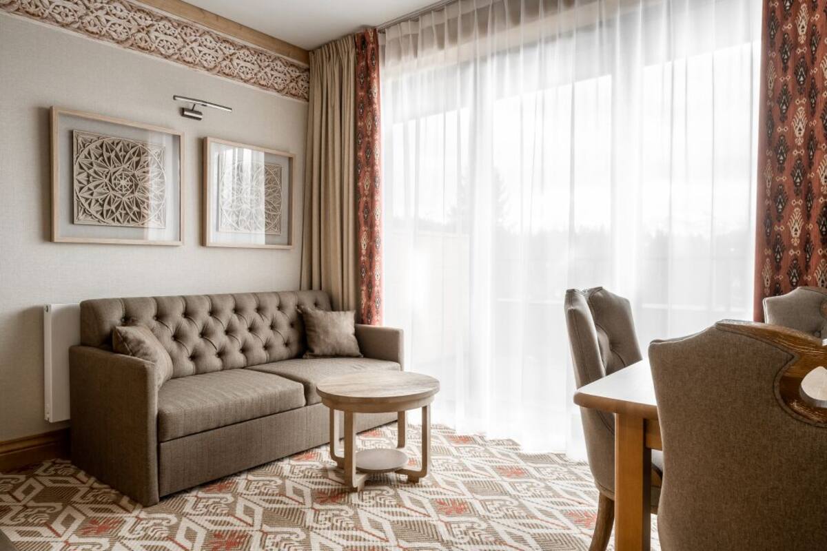 Un salon comprenant un canapé, une table basse ronde et une table à manger avec des chaises. On y trouve deux éléments de décoration encadrés accrochés au mur, ainsi qu’une fenêtre ornée de rideaux légers.