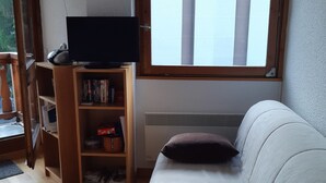 Un salon avec un canapé blanc, une bibliothèque en bois, un téléviseur à écran plat et une fenêtre équipée de stores.