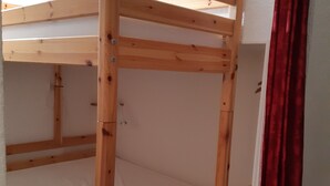 Un lit superposé avec une structure en bois et des vis en métal, un matelas blanc et un rideau rouge, dont on ne voit qu’une partie.