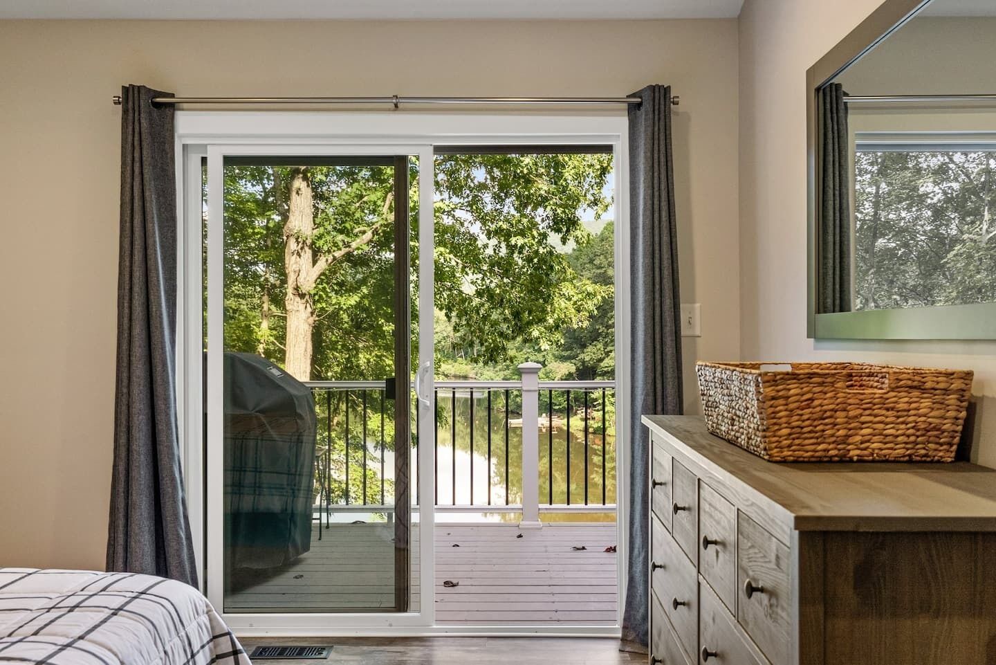 A room with a bed, a wooden dresser, a basket, and a sliding glass door leading to a balcony with a view of trees.