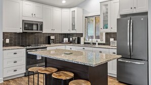 Eine moderne Küche mit weißen Schränken, einer Granitinsel mit Barhocker, einem Edelstahlkühlschrank und einer Mikrowelle.